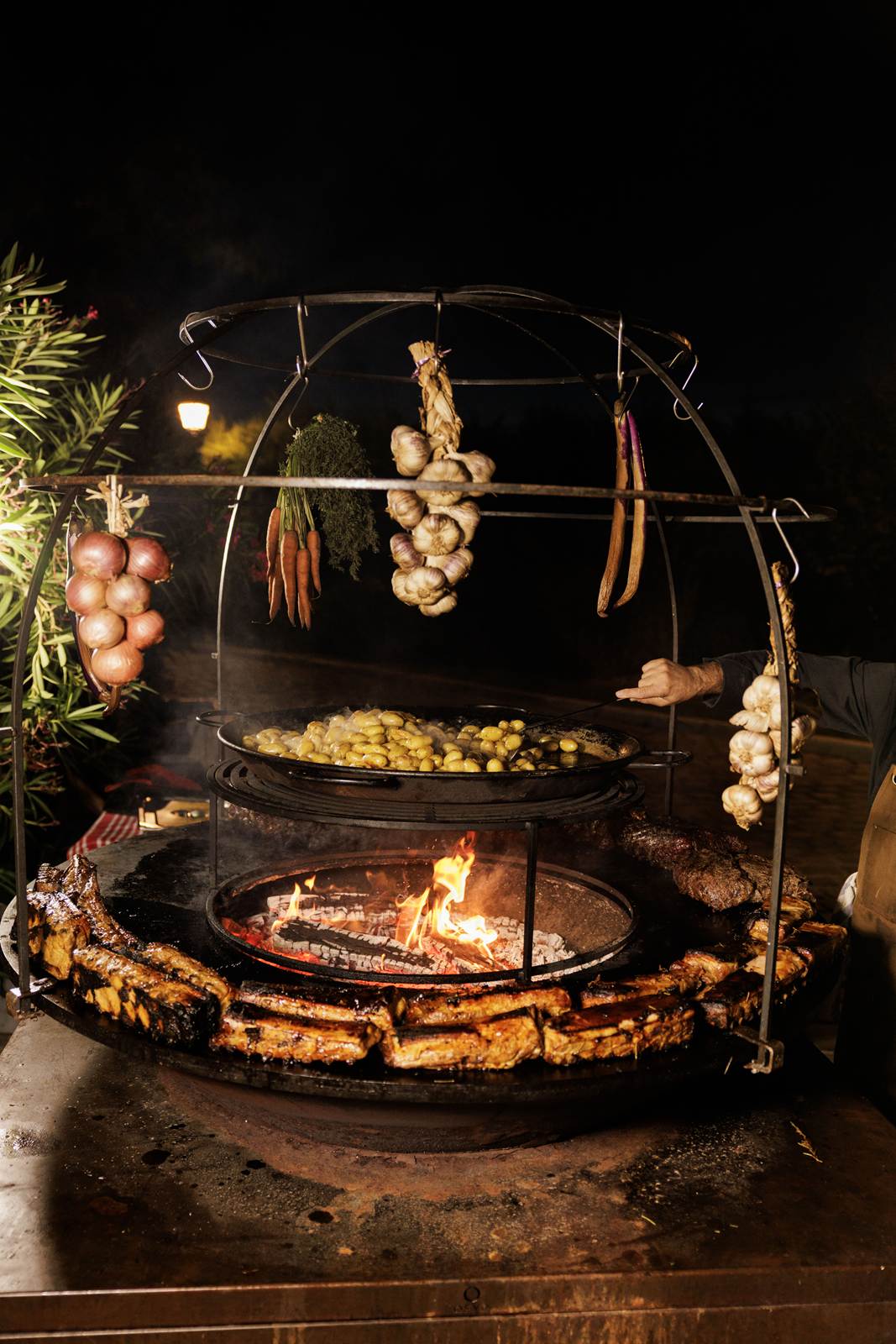 A brasero at night, beneath braids of garlic, onions and carrots