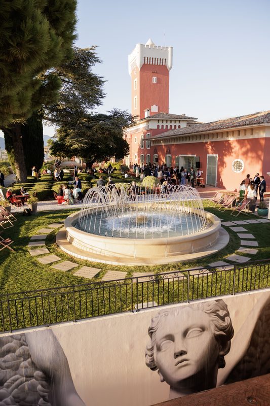 A cocktail party in the gardens of a villa with a tower, around a fountain